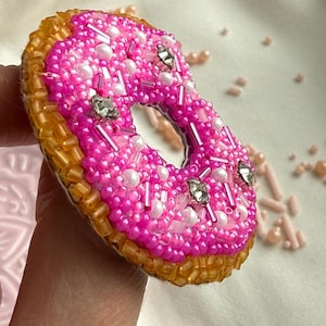 Puede incluir: Un broche con forma de d&oacute;nut elaborado con cuentas de semillas de color rosa vibrante, cuentas blancas tipo perla y detalles de cristal transparente. El anillo exterior est&aacute; cubierto de cuentas tubulares doradas, creando una textura de pastel.