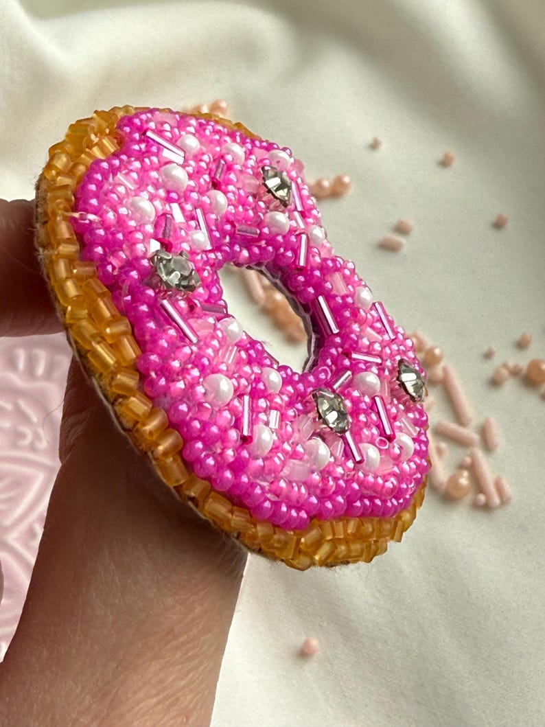 Puede incluir: Un broche con forma de d&oacute;nut elaborado con cuentas de semillas de color rosa vibrante, cuentas blancas tipo perla y detalles de cristal transparente. El anillo exterior est&aacute; cubierto de cuentas tubulares doradas, creando una textura de pastel.