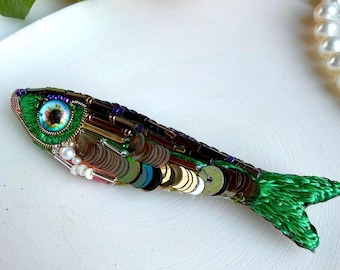 Puede incluir: Un broche con forma de pez elaborado con hilo de bordar verde, con un ojo prominente de iris azul y pupila marrón. El cuerpo está adornado con lentejuelas metálicas marrones, doradas y plateadas, junto con cuentas de vidrio moradas y marrones.