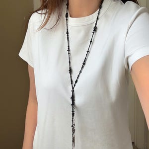 Puede incluir: Un collar largo de cuentas negras con un diseño en forma de Y. El collar presenta pequeñas cuentas oscuras y un colgante colgante con un dije ovalado plateado. El collar se usa sobre una camiseta blanca.