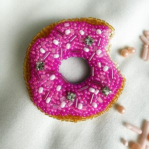 Puede incluir: Un broche de donut hecho con cuentas que presenta una superficie de glaseado rosa vibrante decorada con cuentas blancas y rosas, cristales transparentes y un borde dorado. La forma del donut tiene un mordisco en el lado derecho.
