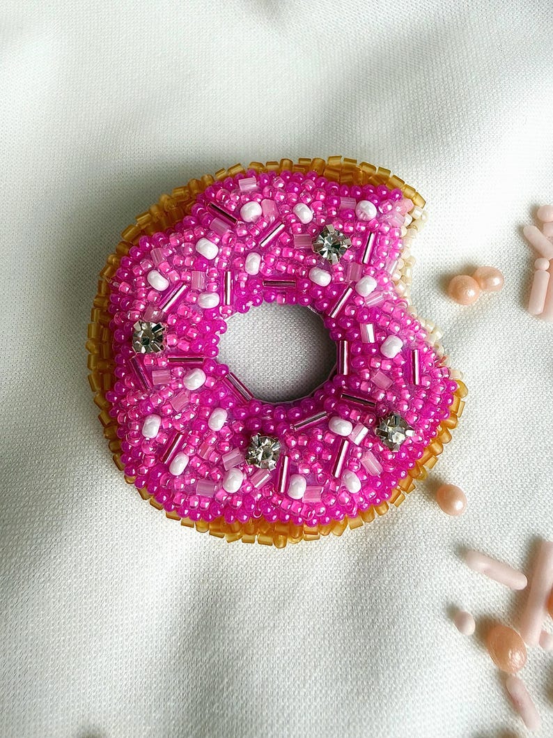 Puede incluir: Un broche de donut hecho con cuentas que presenta una superficie de glaseado rosa vibrante decorada con cuentas blancas y rosas, cristales transparentes y un borde dorado. La forma del donut tiene un mordisco en el lado derecho.