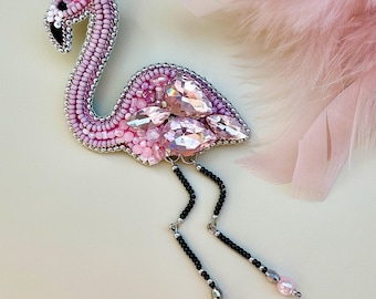 Puede incluir: Un broche con forma de flamenco hecho con cuentas de semillas rosas y blancas, con grandes cristales facetados de color rosa claro en el cuerpo. El ave tiene un ojo negro y dos patas colgantes de cuentas negras con detalles rosas y plateados.