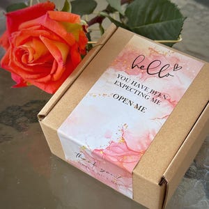 Puede incluir: Una caja cuadrada de cart&oacute;n marr&oacute;n con una etiqueta decorativa de dise&ntilde;o de acuarela rosa y blanco, motas doradas y el texto: hello, YOU HAVE BEEN EXPECTING ME, OPEN ME, thank you, HAVE A NICE DAY. Una rosa naranja y amarilla vibrante est&aacute; al lado.