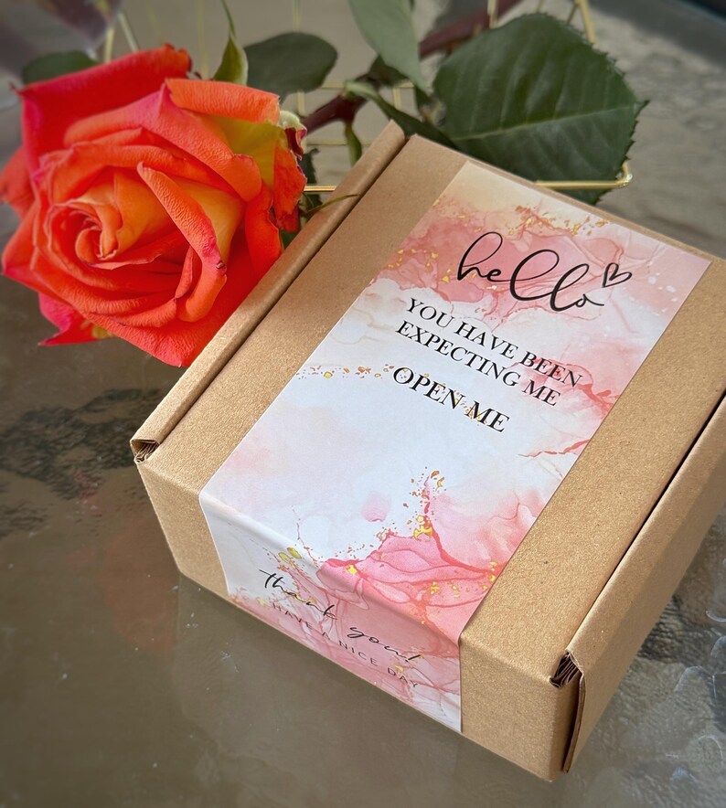 Puede incluir: Una caja cuadrada de cart&oacute;n marr&oacute;n con una etiqueta decorativa de dise&ntilde;o de acuarela rosa y blanco, motas doradas y el texto: hello, YOU HAVE BEEN EXPECTING ME, OPEN ME, thank you, HAVE A NICE DAY. Una rosa naranja y amarilla vibrante est&aacute; al lado.
