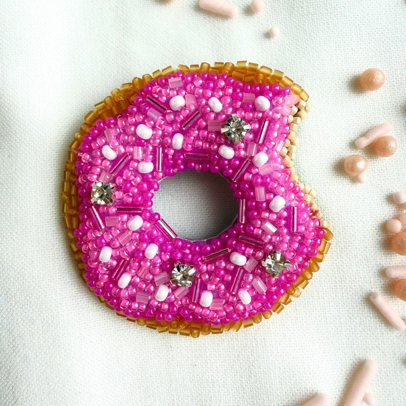Puede incluir: Un broche con forma de d&oacute;nut elaborado con cuentas de semillas de color rosa vibrante, cuentas blancas y cristales transparentes. El borde exterior est&aacute; forrado con cuentas doradas met&aacute;licas y el dise&ntilde;o presenta un peque&ntilde;o mordisco a la derecha.