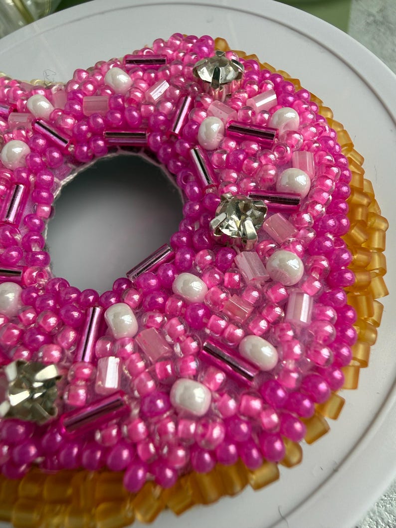 Puede incluir: Un adorno con forma de d&oacute;nut cubierto de cuentas de semillas de color rosa vibrante, cuentas tubulares de color rosa transl&uacute;cido y cuentas blancas nacaradas. El dise&ntilde;o presenta cristales facetados transparentes y un borde de cuentas color &aacute;mbar.