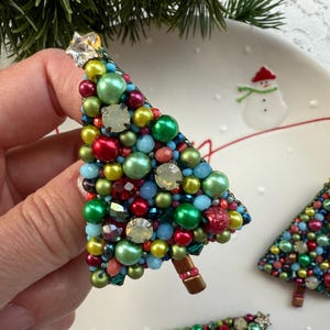 Puede incluir: Un broche de árbol de Navidad, adornado con cuentas de colores en tonos verdes, rojos, amarillos y azules. El árbol está rematado con un adorno en forma de estrella. El broche tiene un tronco marrón y está sobre un fondo blanco.
