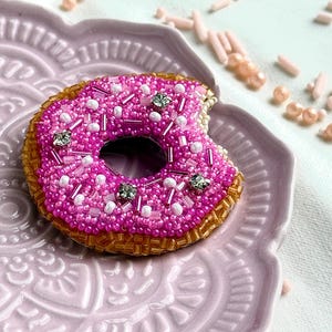Puede incluir: Un broche de donut con cuentas que presenta una cobertura rosa brillante con detalles en blanco y cristales transparentes, sobre una base de color marr&oacute;n dorado. La forma del donut incluye una marca de mordisco en el lado derecho, hecha con cuentas.