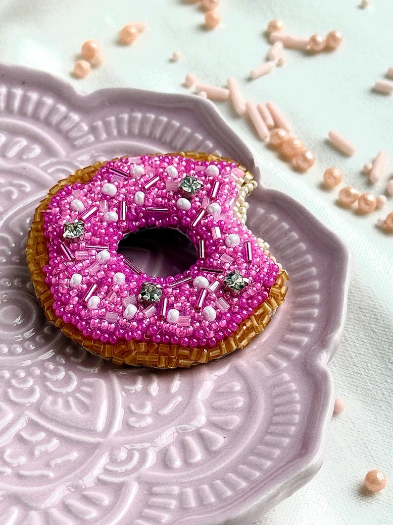 Puede incluir: Un broche de donut con cuentas que presenta una cobertura rosa brillante con detalles en blanco y cristales transparentes, sobre una base de color marr&oacute;n dorado. La forma del donut incluye una marca de mordisco en el lado derecho, hecha con cuentas.