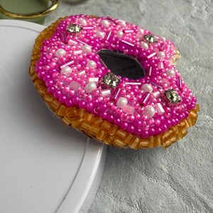 Puede incluir: Un broche con forma de d&oacute;nut decorado con cuentas de color rosa vibrante, detalles blancos nacarados y cristales transparentes en la parte superior, con un borde de cuentas de color &aacute;mbar transl&uacute;cido que forma la base del d&oacute;nut.