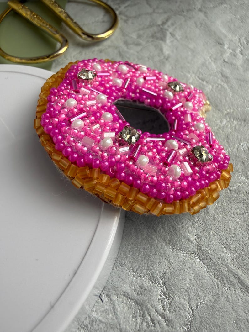 Puede incluir: Un broche con forma de d&oacute;nut decorado con cuentas de color rosa vibrante, detalles blancos nacarados y cristales transparentes en la parte superior, con un borde de cuentas de color &aacute;mbar transl&uacute;cido que forma la base del d&oacute;nut.