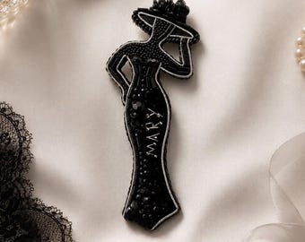 Puede incluir: Un broche de cuentas negras con la silueta de una persona con sombrero de ala ancha y vestido largo. El vestido lleva el nombre MARY bordado en hilo plateado, acentuado con varias cuentas negras y un borde plateado.