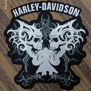 May include: Black and white embroidered patch featuring a Harley-Davidson logo and two skulls with flames. The skulls are surrounded by intricate floral designs.