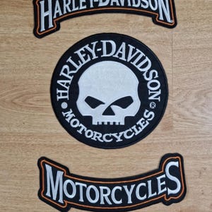 Könnte beinhalten: Drei Harley-Davidson Motorrad-Patches. Der obere Patch ist ein schwarzes und oranges Banner mit dem Text "Harley-Davidson". Der mittlere Patch ist ein schwarzer Kreis mit einem weißen Totenkopf und gekreuzten Knochen und dem Text "Harley-Davidson Motorcycles". Der untere Patch ist ein schwarzes und oranges Banner mit dem Text "Motorcycles".