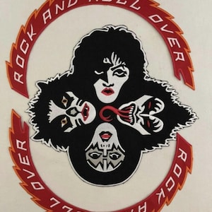 Może przedstawiać: Naszywkę haftowaną w czerni i bieli przedstawiającą czterech członków zespołu rockowego KISS. Naszywka jest otoczona czerwono-pomarańczowym kółkiem z napisem "Rock and Roll Over".