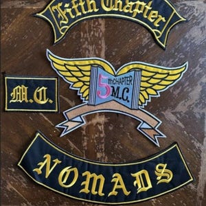 Può includere: Cinque toppe ricamate in nero e oro con le parole "Fifth Chapter", "M.C.", "5th Chapter M.C." e "NOMADS". Le toppe mostrano un libro con le ali e uno stendardo.