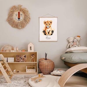 May include: A nursery room with a light brown wooden shelf with a toy camera, wooden blocks, and a wooden toy house. There is a light brown wicker basket, a light brown wooden rocker, and a white rug with a light brown rug underneath. A framed print of a lion cub with the name Leon is hanging on the wall. A light brown woven lion head is hanging on the wall.