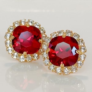May include: A pair of gold-toned stud earrings. Each earring features a large, square, faceted, ruby-red gemstone surrounded by a halo of smaller, clear, round gemstones. The earrings are set against a white background.