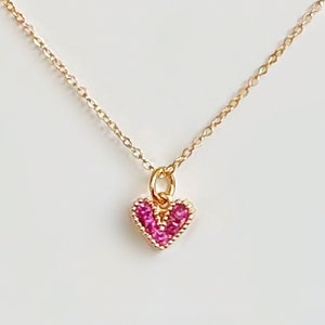 Può includere: Una delicata collana a catena dorata con un piccolo ciondolo a forma di cuore rosa. Il cuore è adornato con minuscole pietre preziose rosa.