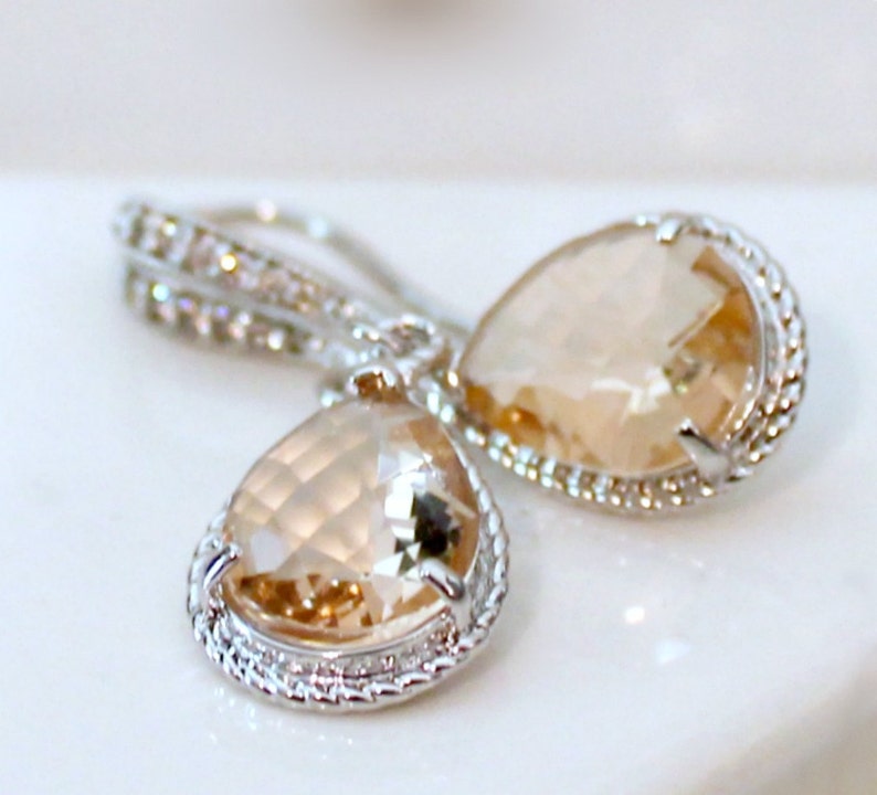May include: A pair of silver teardrop earrings with clear, faceted, champagne-colored stones. The stones are set in a silver bezel with a decorative, rope-like border.