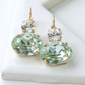 May include: A pair of gold-toned earrings featuring a clear, round crystal above a large, oval, light green gemstone. The earrings have a lever-back closure and are set on a white surface.