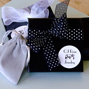 May include: A black gift box with a white polka dot ribbon and a white circular label that says "CJ Rose Jewelry" with a cartoon rhinoceros illustration. The box is sitting on a light blue surface with a gray velvet bag.