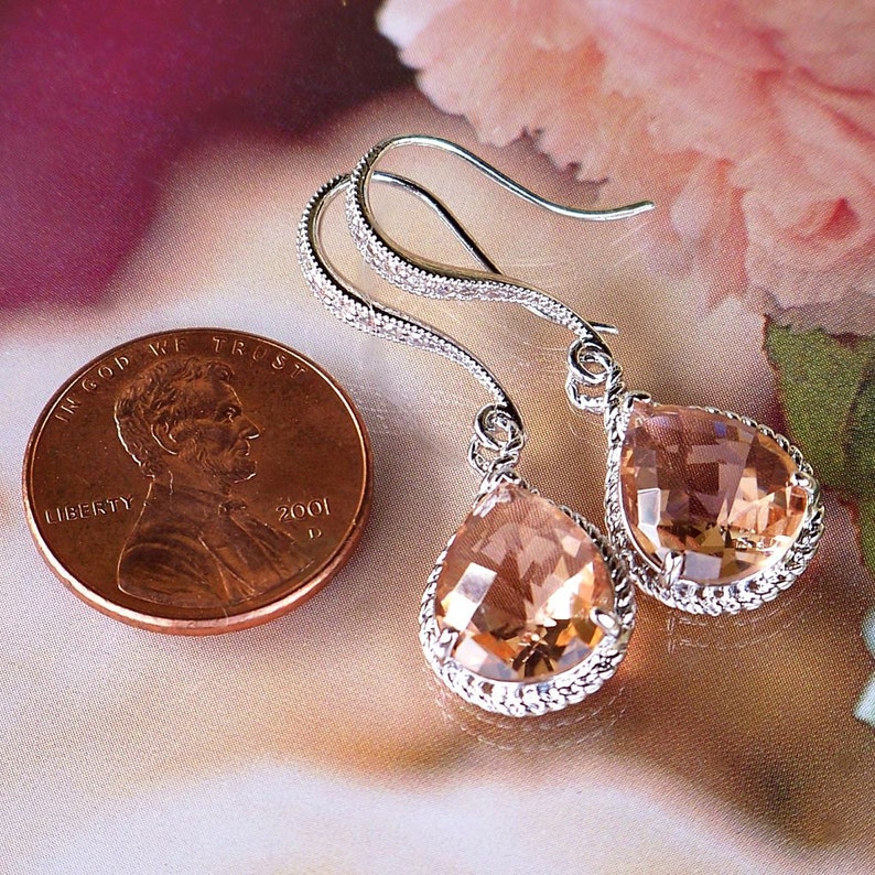 May include: A pair of silver teardrop earrings with a peach-colored crystal. The earrings have a delicate, sparkly design.