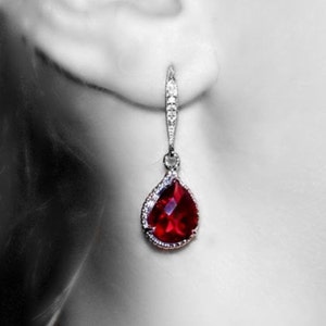 May include: A pair of dangle earrings with a teardrop-shaped red gemstone surrounded by smaller clear gemstones. The earrings are silver in color.