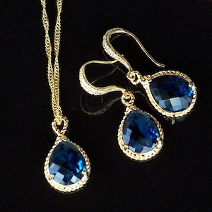 May include: A gold necklace and earrings set with teardrop-shaped blue gemstone pendants. The pendants have a rope-like gold trim.