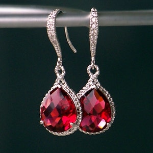 Puede incluir: Un par de pendientes de plata en forma de lágrima con piedras de cristal rojas. Los pendientes tienen un diseño delicado y retorcido y son perfectos para añadir un toque de brillo a cualquier atuendo.