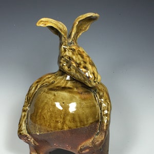 May include: A ceramic sculpture of a rabbit with long ears and a stout body. The rabbit is glazed in shades of brown and tan, with a glossy finish. The sculpture has a whimsical, folk-art style.