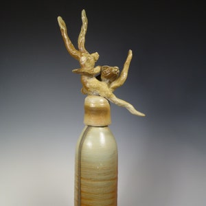 May include: A tall, ceramic bottle with a sculpted, abstract figure on top. The bottle is primarily cream-colored with horizontal textured bands and brown accents. The figure is a light brown color.