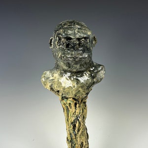 May include: A ceramic sculpture of a gorilla head on a tree trunk with a small black ceramic animal at the base.