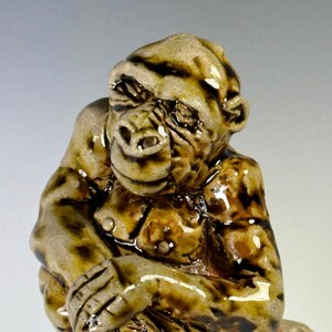 May include: A ceramic sculpture of a brown and tan gorilla sitting with its arms crossed. The gorilla is glazed and has a detailed face.