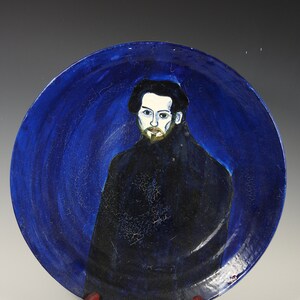 May include: A ceramic plate with a portrait of a man. The plate has a deep blue background with a crackle effect. The man is depicted with a black coat, black hat, and a light complexion with a beard. The plate is displayed on a red stand.