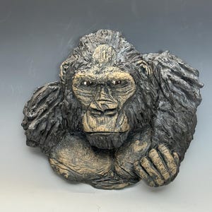 May include: A detailed sculpture of a gorilla's head and upper body. The gorilla is depicted with a stern expression, featuring a textured, dark gray fur and a lighter tan face. The sculpture is a wall hanging, showcasing the gorilla's powerful physique.