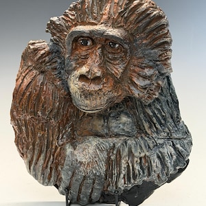 May include: A detailed ceramic sculpture of a primate's head and upper body. The sculpture features a textured surface with shades of brown, gray, and black. The primate's face has a contemplative expression, and the sculpture is mounted on a black stand.