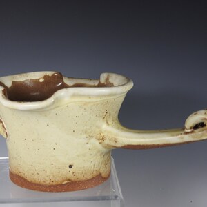 May include: A handmade ceramic gravy boat with a unique, asymmetrical design. The boat has a light yellow glaze with brown speckles and a brown rim. It has a single handle that curves up and ends in a swirl.