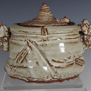 May include: A ceramic jar with a lid, featuring a textured, white glaze and brown accents. The jar has two sculpted handles in the shape of faces.