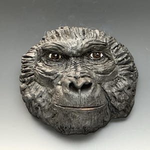 May include: A detailed, gray sculpture of a gorilla's face. The sculpture features realistic textures, brown eyes, and a subtle smile. The artwork is a wall hanging, showcasing the primate's features.