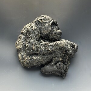 May include: A detailed sculpture of a chimpanzee in a seated position, rendered in shades of gray and black. The ape's fur appears textured, with visible details in its face and limbs. The sculpture is set against a gray background.
