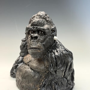 May include: A dark charcoal-colored gorilla bust sculpture featuring a textured, rugged surface that mimics fur. The gorilla has a prominent brow, deep-set eyes, and a small, singular white circular accent attached to the side of its head.
