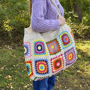 May include: A large, cream-colored crochet tote bag with colorful square patterns. The squares feature vibrant hues of pink, yellow, blue, and orange. The bag is carried over the shoulder.