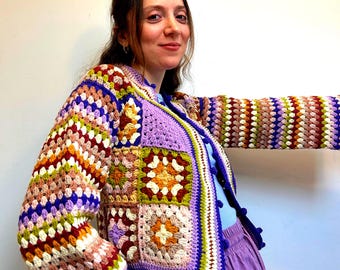 May include: A colorful crocheted cardigan with a patchwork design featuring squares in shades of purple, green, orange, and brown. The cardigan has a button closure and a crocheted trim.