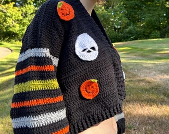 May include: A black crochet cardigan featuring long sleeves with horizontal stripes in orange, lime green, and light gray. The front of the garment is decorated with crocheted appliques including two orange pumpkins with green stems and a white skull.