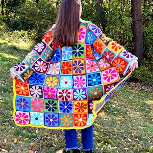 May include: A colorful crochet cardigan with a floral pattern. The cardigan is made up of squares with different colored flowers in the center. The cardigan is worn by a person with long brown hair.