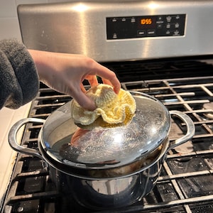 May include: A stainless steel pot with a lid, on a black stovetop. A yellow crocheted item is on top of the lid. The hand of a person is holding the crocheted item. The time 10:55 is displayed on the oven.