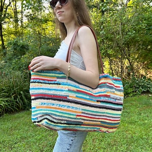 May include: A large, colorful crochet tote bag with a striped pattern in various colors, including blue, red, yellow, and green. The bag has brown leather handles and a rectangular shape. The bag is held by a person wearing sunglasses and blue jeans.