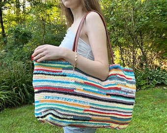 May include: A large, colorful crochet tote bag with a striped pattern in various colors, including blue, red, yellow, and green. The bag has brown leather handles and a rectangular shape. The bag is held by a person wearing sunglasses and blue jeans.